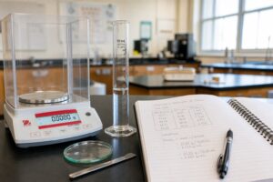 Un banc de laboratoire de physique-chimie avec une balance numérique, une éprouvette graduée et un cahier d'étudiant contenant des données de mesure, le tout éclairé par une lumière naturelle tamisée.